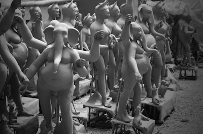 The image depicts multiple clay statues in various stages of completion arranged closely together. One prominent statue in the foreground features an anthropomorphic elephant figure. Other statues display humanlike forms, some with humanoid faces while others have animalistic features. The statues are placed on individual platforms.