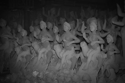A collection of clay sculptures depicting figures in various poses. These figures appear to be in the process of being molded and finished. They are arranged closely together, creating a dense visual effect. Some sculptures are more detailed than others, featuring defined facial features and gestures. The environment appears to be a workshop or storage space for these unfinished sculptures.