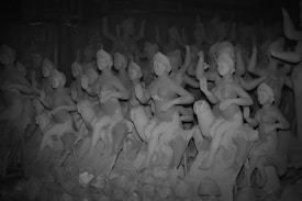 A collection of clay sculptures depicting figures in various poses. These figures appear to be in the process of being molded and finished. They are arranged closely together, creating a dense visual effect. Some sculptures are more detailed than others, featuring defined facial features and gestures. The environment appears to be a workshop or storage space for these unfinished sculptures.