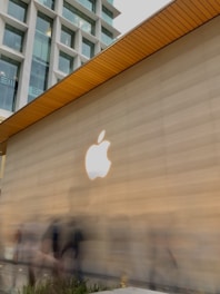 A large beige wall features a prominent white Apple logo, with a modern building visible in the background. The ceiling has a wooden texture with a series of lines. People are walking by, appearing as blurred figures due to motion.