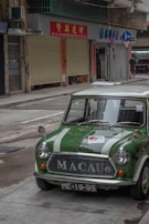 A vintage green car with white racing stripes is parked on a street. The car has 'MACAU' written on its grille and the license plate reads 'ME-19-95'. The surroundings include commercial buildings with shuttered shopfronts, a man walking in the distance, and directional signage.