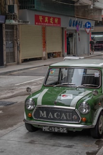 A vintage green car with white racing stripes is parked on a street. The car has 'MACAU' written on its grille and the license plate reads 'ME-19-95'. The surroundings include commercial buildings with shuttered shopfronts, a man walking in the distance, and directional signage.