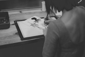A person is engaged in drawing on a piece of paper placed on a table. The drawing appears intricate, featuring an artistic design with organic and possibly tribal elements. The scene is captured in black and white, which adds a classic and focused aesthetic to the workspace.