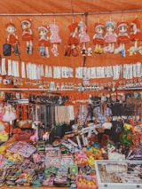 A vibrant market stall showcasing a wide variety of small goods and toys. There are several plush dolls in red outfits hanging at the top, alongside various keychains and necklaces. Below, the stall is filled with an assortment of colorful items, including toy guns, action figures, and other trinkets. The display is busy and visually engaging, with items densely packed.