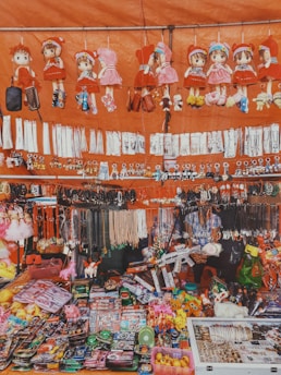 A vibrant market stall showcasing a wide variety of small goods and toys. There are several plush dolls in red outfits hanging at the top, alongside various keychains and necklaces. Below, the stall is filled with an assortment of colorful items, including toy guns, action figures, and other trinkets. The display is busy and visually engaging, with items densely packed.