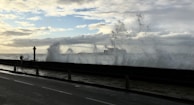 Large ocean waves crash against a seawall, sending sprays of water into the air. A person stands on the promenade, possibly taking a photo or video with a mobile device. In the background, the sky is partly cloudy with patches of blue visible.
