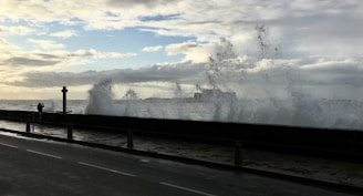Large ocean waves crash against a seawall, sending sprays of water into the air. A person stands on the promenade, possibly taking a photo or video with a mobile device. In the background, the sky is partly cloudy with patches of blue visible.