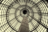A detailed view of an architectural structure featuring a large, circular glass and metal dome that rises above a brick tower. The intricate pattern of beams forms a complex geometric design converging at the dome's center, creating a sense of depth.
