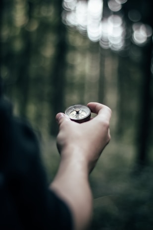 A person holds a compass in their hand against a blurred background of a forest, indicating a setting of exploration or navigation.