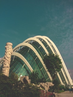 A modern glass structure with curved arches stands against a blue sky, surrounded by lush greenery and a tall sculpted column. The architectural design features large transparent panels reflecting the sunlight, complemented by landscaped rocks and trees.