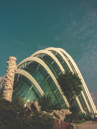 A modern glass structure with curved arches stands against a blue sky, surrounded by lush greenery and a tall sculpted column. The architectural design features large transparent panels reflecting the sunlight, complemented by landscaped rocks and trees.