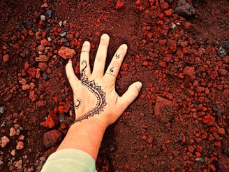 A hand with intricate henna designs is placed on a surface covered with small, red gravel-like stones. The henna pattern is detailed with swirling shapes and triangular motifs, contrasting with the earthy background.