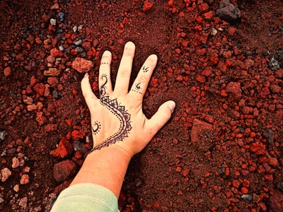 A hand with intricate henna designs is placed on a surface covered with small, red gravel-like stones. The henna pattern is detailed with swirling shapes and triangular motifs, contrasting with the earthy background.