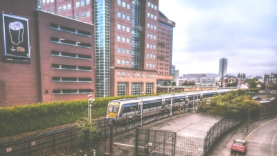 A commuter train travels alongside a large office building with red brick facades and a prominent poster of a beverage on the side. The scene includes a blend of urban elements like industrial fences, a road with a few parked cars, and distant cityscape buildings under a cloudy sky.