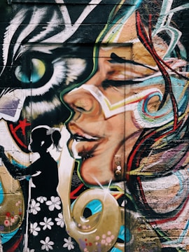 A vibrant and detailed mural painted on a wall. The artwork features a large, colorful face with abstract designs and flowing lines. In the foreground, there's a black silhouette of a girl wearing a dress with flower patterns. An eye that resembles that of a bird or a cat is also part of the mural, giving it a mystical vibe.