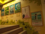 A staircase scene with framed anime movie posters mounted on a wall. The posters include colorful illustrations and Japanese text. The wall is decorated with green leaves and cartoon-like creatures, enhancing a playful and artistic ambiance.