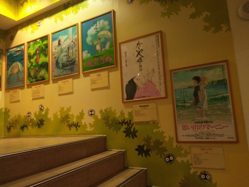 A staircase scene with framed anime movie posters mounted on a wall. The posters include colorful illustrations and Japanese text. The wall is decorated with green leaves and cartoon-like creatures, enhancing a playful and artistic ambiance.