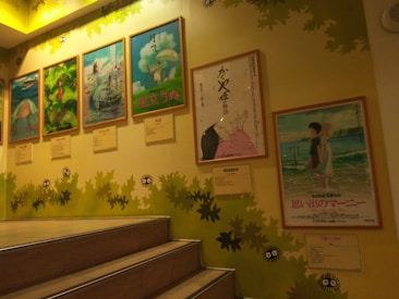 A staircase scene with framed anime movie posters mounted on a wall. The posters include colorful illustrations and Japanese text. The wall is decorated with green leaves and cartoon-like creatures, enhancing a playful and artistic ambiance.