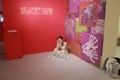 A woman in a floral dress is sitting on the floor in a gallery room with red walls. The space is part of a solo exhibition by Ban-Yuan Chang. The wall features art panels with red, pink, and green backgrounds covered in cryptic, digital-like text and images of hands.