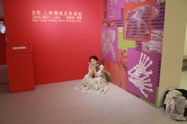 A woman in a floral dress is sitting on the floor in a gallery room with red walls. The space is part of a solo exhibition by Ban-Yuan Chang. The wall features art panels with red, pink, and green backgrounds covered in cryptic, digital-like text and images of hands.