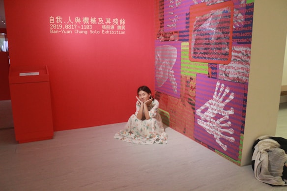 A woman in a floral dress is sitting on the floor in a gallery room with red walls. The space is part of a solo exhibition by Ban-Yuan Chang. The wall features art panels with red, pink, and green backgrounds covered in cryptic, digital-like text and images of hands.