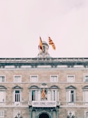 A historic building features multiple flags, including Spanish and Catalan flags, prominently displayed on its rooftop and facade. The building is made of stone with symmetrical windows and classic architectural details. A large banner hangs over the entrance, reading 'Llibertat d'Opinió i d'Expressió.'