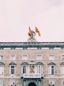 A historic building features multiple flags, including Spanish and Catalan flags, prominently displayed on its rooftop and facade. The building is made of stone with symmetrical windows and classic architectural details. A large banner hangs over the entrance, reading 'Llibertat d'Opini&oacute; i d'Expressi&oacute;.'
