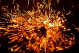 Fiery sparks in vivid orange burst outward, creating a dynamic and intense visual effect. Glowing specks and streaks light up against a dark background, resembling an explosion or a sparkler. The image captures the motion and energy of the particles as they scatter.