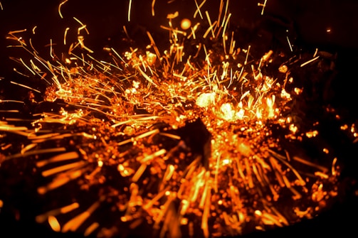 Fiery sparks in vivid orange burst outward, creating a dynamic and intense visual effect. Glowing specks and streaks light up against a dark background, resembling an explosion or a sparkler. The image captures the motion and energy of the particles as they scatter.