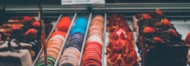 An assortment of colorful macarons is lined up in a display case alongside various chocolate pastries topped with strawberries. The display is well-lit, showcasing the vibrant colors of the macarons in shades of orange, pink, purple, and blue, as well as the rich, dark tones of the chocolate desserts.