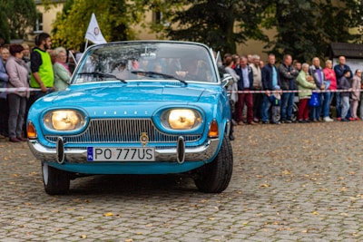 A vintage blue car is parked on a cobblestone street with headlights on, surrounded by a crowd of people. The car's number plate reads 'PO 777US' and a small flag is attached near the side mirror. Many of the onlookers are standing behind a striped barrier, and the setting is outdoors with trees and buildings in the background.