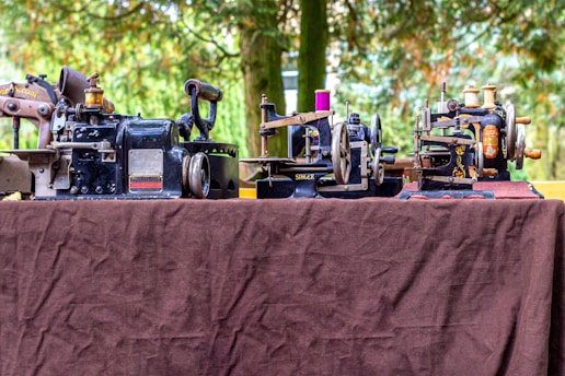 A collection of vintage sewing machines is displayed on a brown cloth-covered table outdoors. There are various styles and sizes, mostly in black with metallic components, each having intricate designs. The background features a lush green setting with blurred greenery, suggesting a park or garden.