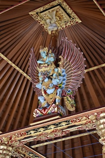 A vibrant, intricately carved statue of a mythological figure with blue skin and ornate golden armor. The figure has large, colorful wings and is positioned under a pointed, decorative wooden ceiling with detailed gold trim.
