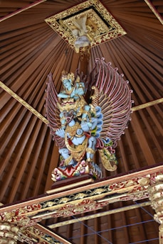 A vibrant, intricately carved statue of a mythological figure with blue skin and ornate golden armor. The figure has large, colorful wings and is positioned under a pointed, decorative wooden ceiling with detailed gold trim.