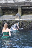 Two individuals are performing a ritual under a stone water fountain, surrounded by moss-covered stone walls. The setting appears to be a traditional or historical site, with water flowing from a carved spout.