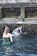 Two individuals are performing a ritual under a stone water fountain, surrounded by moss-covered stone walls. The setting appears to be a traditional or historical site, with water flowing from a carved spout.