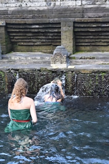 Two individuals are performing a ritual under a stone water fountain, surrounded by moss-covered stone walls. The setting appears to be a traditional or historical site, with water flowing from a carved spout.