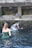 Two individuals are performing a ritual under a stone water fountain, surrounded by moss-covered stone walls. The setting appears to be a traditional or historical site, with water flowing from a carved spout.