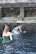 Two individuals are performing a ritual under a stone water fountain, surrounded by moss-covered stone walls. The setting appears to be a traditional or historical site, with water flowing from a carved spout.
