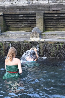 Two individuals are performing a ritual under a stone water fountain, surrounded by moss-covered stone walls. The setting appears to be a traditional or historical site, with water flowing from a carved spout.