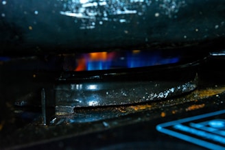 A close-up of a gas stove burner with a blue flame underneath a dark, slightly corroded metal grate. The surrounding area is dark with hints of rust and grime.