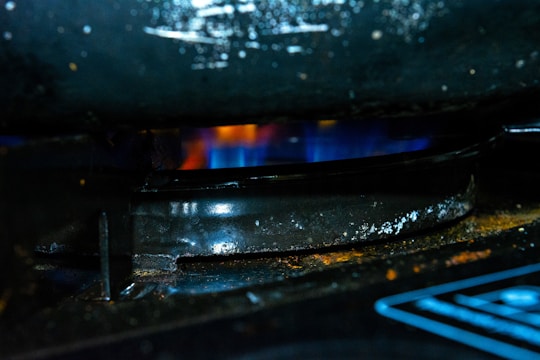 A close-up of a gas stove burner with a blue flame underneath a dark, slightly corroded metal grate. The surrounding area is dark with hints of rust and grime.