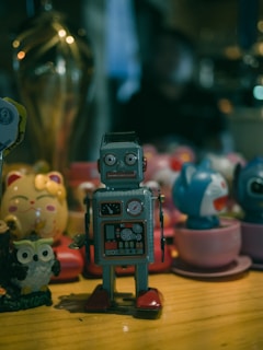 A collection of toys is displayed on a wooden surface, including a vintage-style robot with a square body and red feet, alongside an assortment of colorful figures and decorative items. The background is slightly out of focus, creating a cozy, cluttered atmosphere.