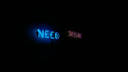 Neon lights in a dark setting spell out the words 'NEED' in blue and 'DESIRE' in purple. The lights illuminate a portion of the surrounding area, creating a stark contrast against the black background.