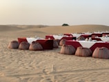 A desert landscape features a rustic outdoor dining setup with tables covered in white and red cloths, accompanied by decorative lanterns and traditional patterned poufs in the sand. The background reveals vast sand dunes with a single bush on the horizon.