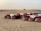 A desert landscape features a rustic outdoor dining setup with tables covered in white and red cloths, accompanied by decorative lanterns and traditional patterned poufs in the sand. The background reveals vast sand dunes with a single bush on the horizon.