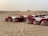 A desert landscape features a rustic outdoor dining setup with tables covered in white and red cloths, accompanied by decorative lanterns and traditional patterned poufs in the sand. The background reveals vast sand dunes with a single bush on the horizon.