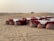A desert landscape features a rustic outdoor dining setup with tables covered in white and red cloths, accompanied by decorative lanterns and traditional patterned poufs in the sand. The background reveals vast sand dunes with a single bush on the horizon.