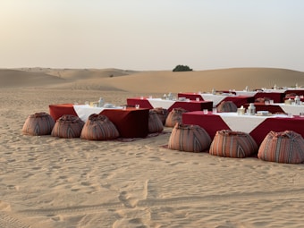 A desert landscape features a rustic outdoor dining setup with tables covered in white and red cloths, accompanied by decorative lanterns and traditional patterned poufs in the sand. The background reveals vast sand dunes with a single bush on the horizon.