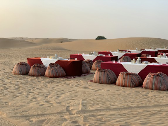 A desert landscape features a rustic outdoor dining setup with tables covered in white and red cloths, accompanied by decorative lanterns and traditional patterned poufs in the sand. The background reveals vast sand dunes with a single bush on the horizon.